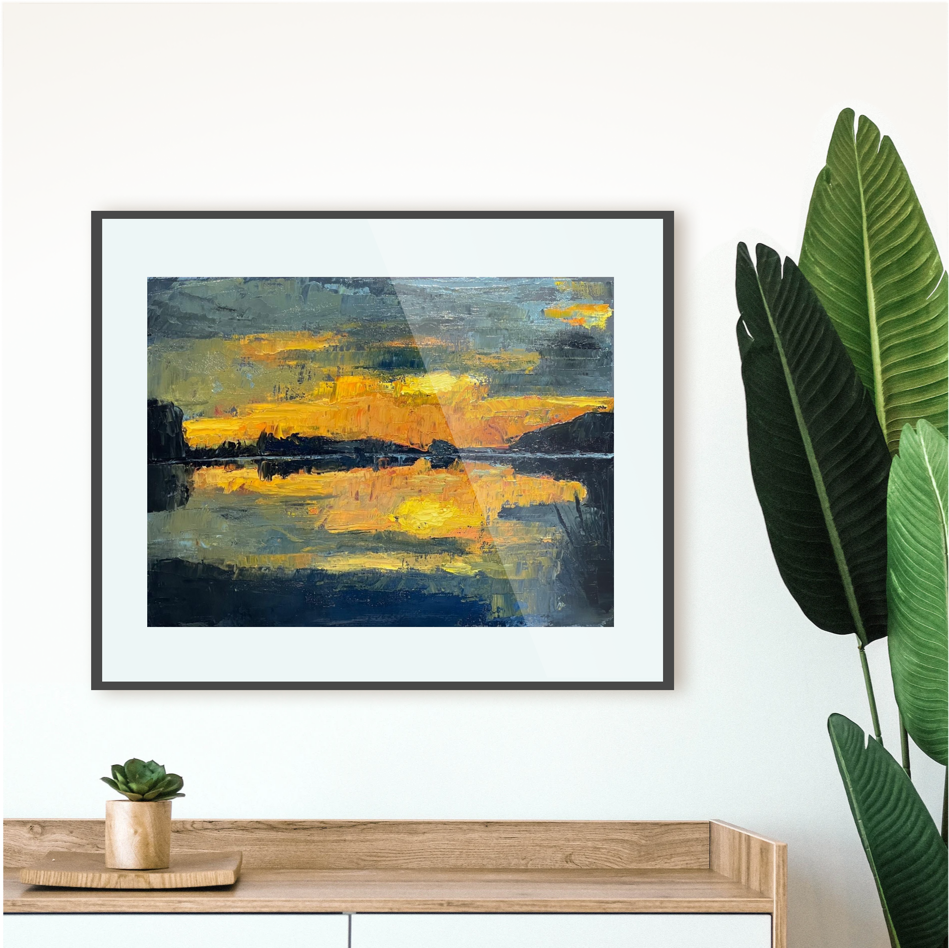Framed abstract artwork with a sunset over water scene, placed above a wooden console table with a plant.