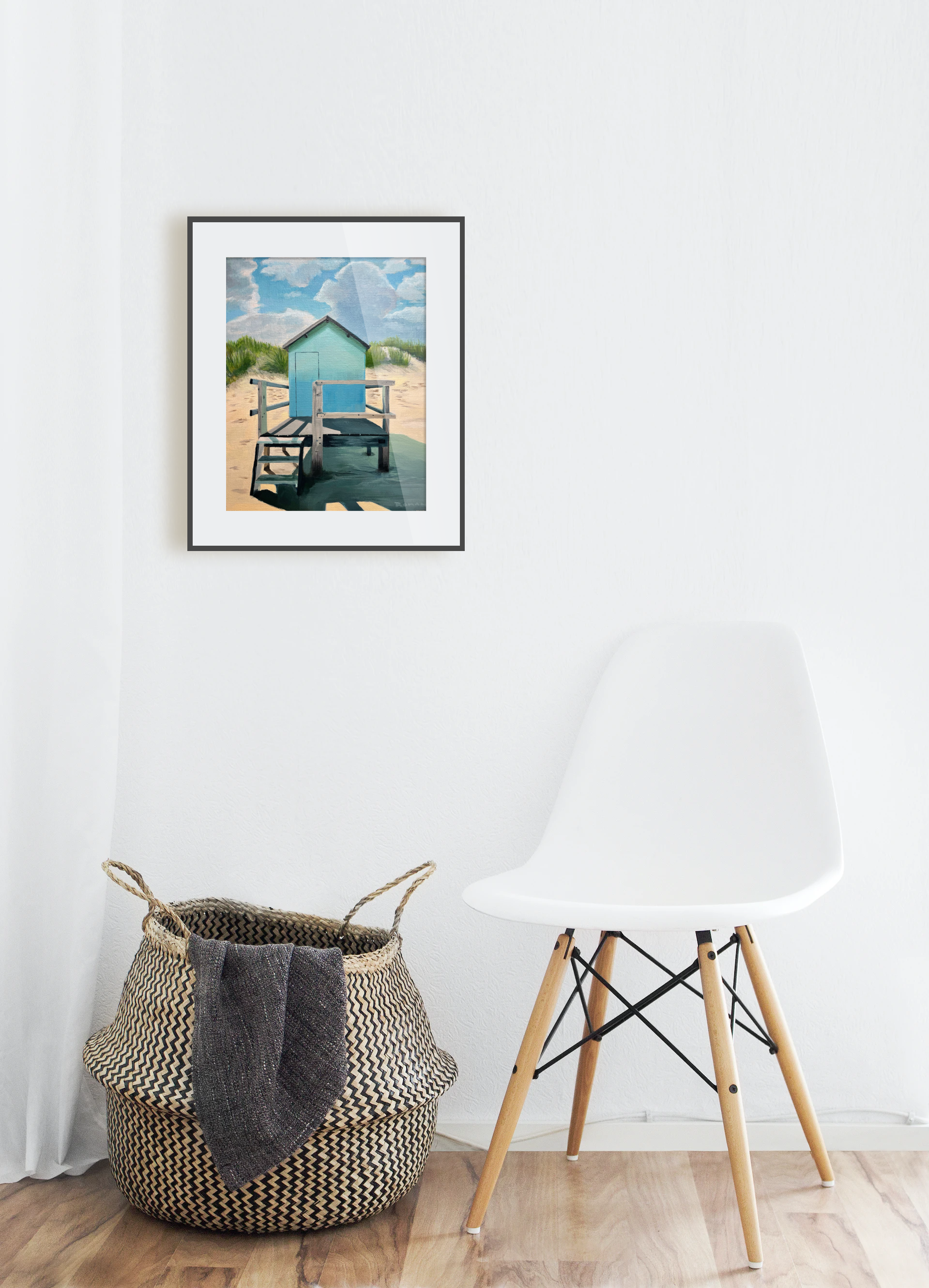 White chair and woven basket against a white wall with a framed picture of a beach scene.